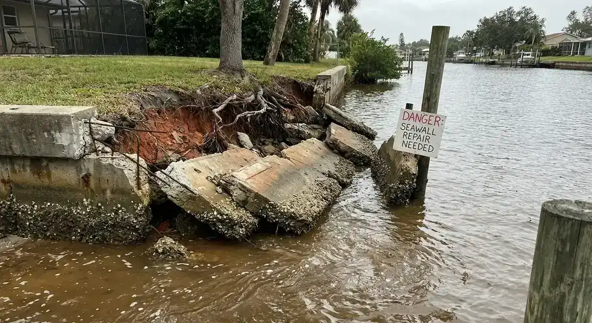 Collapsed concrete seawall panels and eroded yard soil along a residential canal
